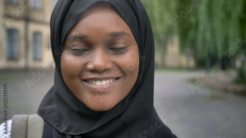Young african american muslim female student in hijab smiling at camera and standing in park near university