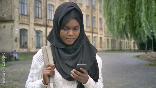 Young beautiful african muslim student in hijab chatting on phone and standing in park near university, holding backpack photo