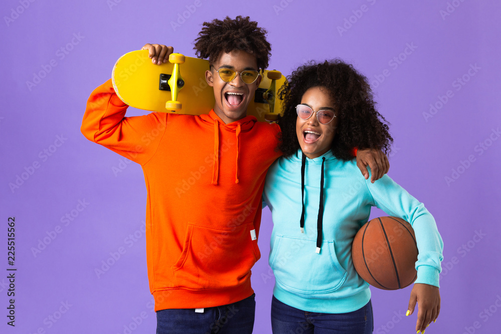 Happy young cute african couple posing isolated over violet background holding skateboard and basketball.