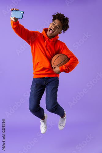 Fototapeta Naklejka Na Ścianę i Meble -  Happy young african man posing isolated over violet background take a selfie by phone holding basketball.