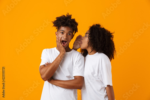 Emotional young cute african couple posing isolated over yellow background gossiping tell secret.