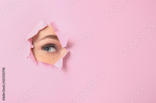Eye of beautiful young woman visible through hole in pink teared paper photo