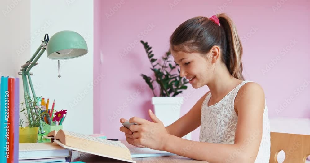 Student teenage girl using cell phone, sitting at the table in bedroom, smiling at camera Student teenage girl using cell phone, sitting at the table in bedroom, smiling at camera