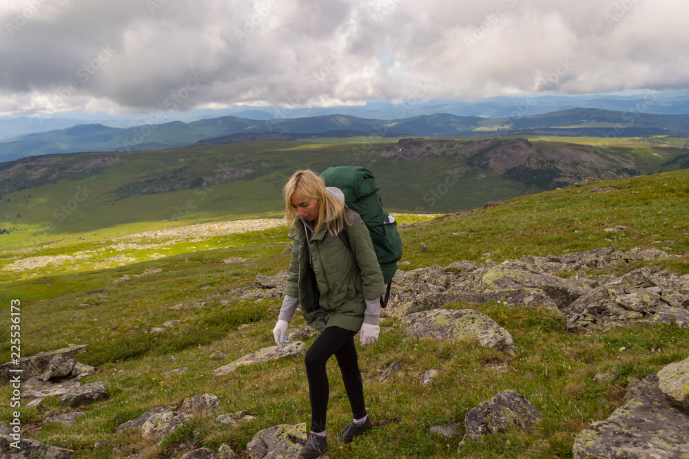 Young beautiful woman travel with big green backpack and bottle, along the green hill with green grass walking on the rocks under the blue sky and big gray and white clouds in Altai mountains