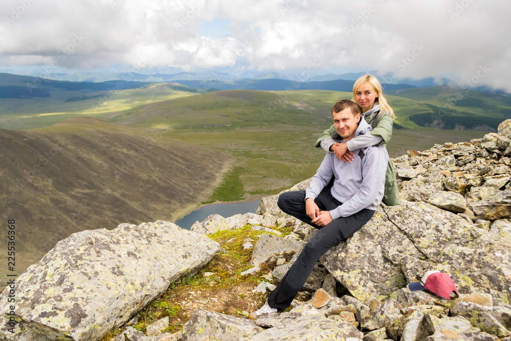 The man sits on a rock, looks into the camera, behind him a blond woman smiles against the background of clouds, blue lake, valley of hills. A loving couple hugs at the top of a mountain in the Altai