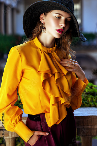  Outdoor fashion portrait of young beautiful woman wearing stylish black wide-brimmed hat, yellow blouse with frills, violet trousers, posing in street, near old european architecture 
