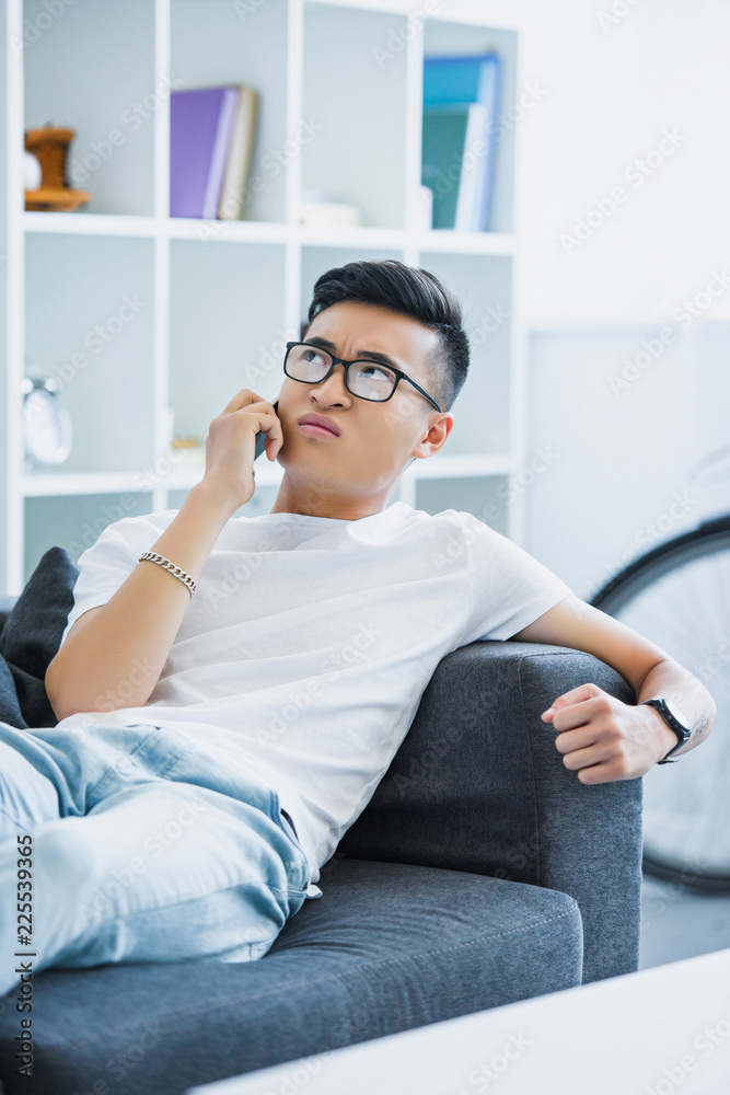 irritated asian man lying on sofa and talking by smartphone at home irritated asian man lying on sofa and talking by smartphone at home