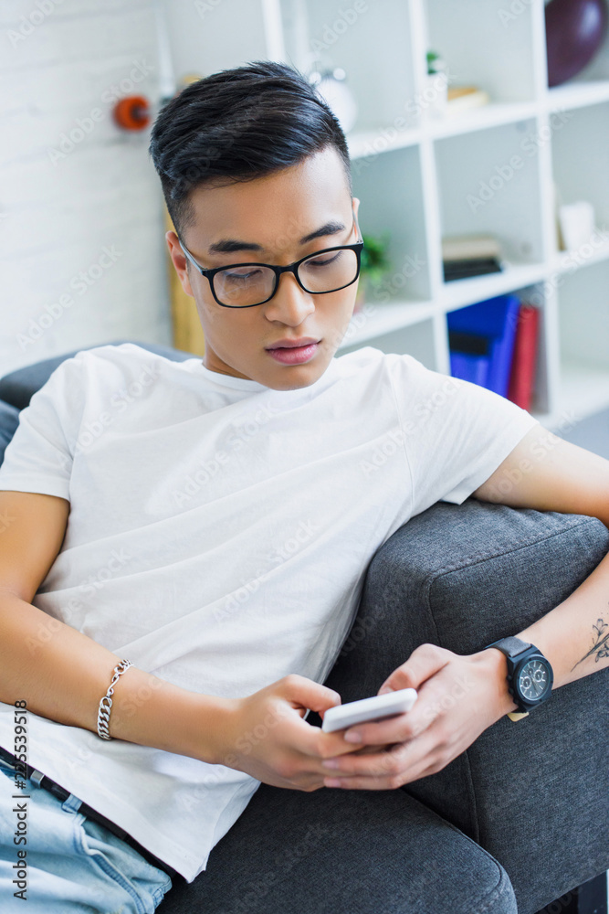 handsome asian man in glasses lying on sofa and using smartphone at home