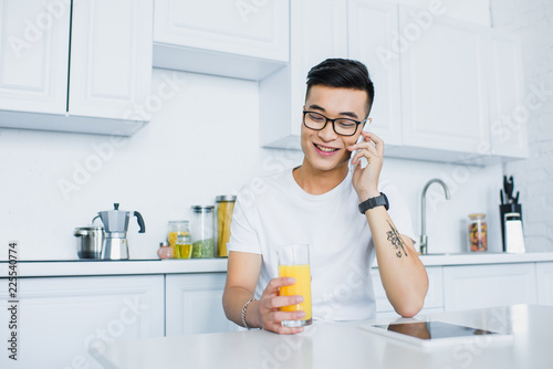 smiling young asian man in eyeglasses holding glass of juice and talking by smartphone while sitting in kitchen
