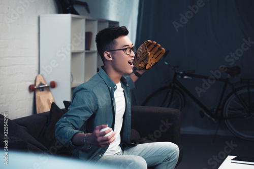 Photography emotional young asian man with baseball glove and ball sitting on couch and watc