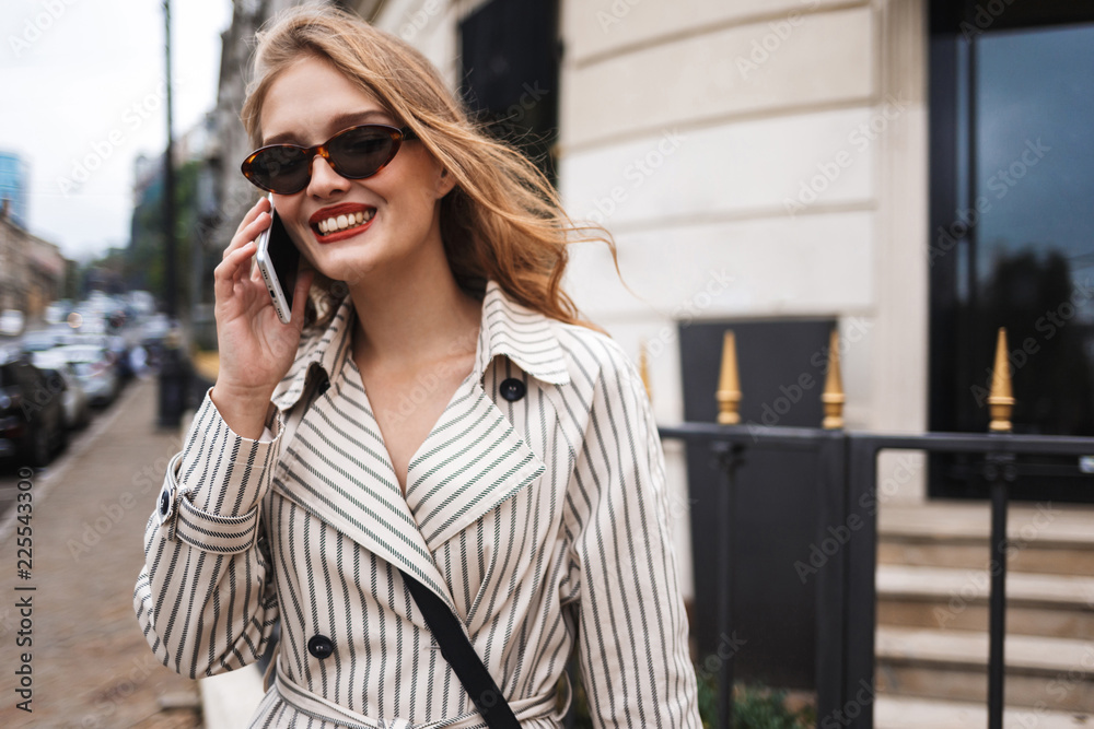 Young pretty smiling woman in striped trench coat happily talking on cellphone while walking around cozy city street