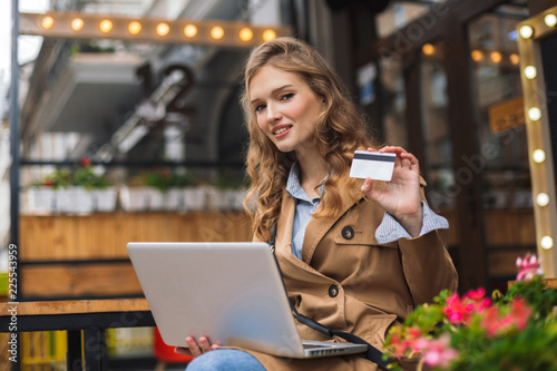Pretty smiling woman in trench coat dreamily looking in camera while holding credit card in hand sitting with laptop on knees outdoor at the cafe terrace