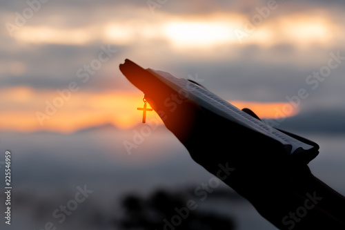 Fotografie, Obraz Silhouette of human hand holding bible and cross, the background is the sunrise