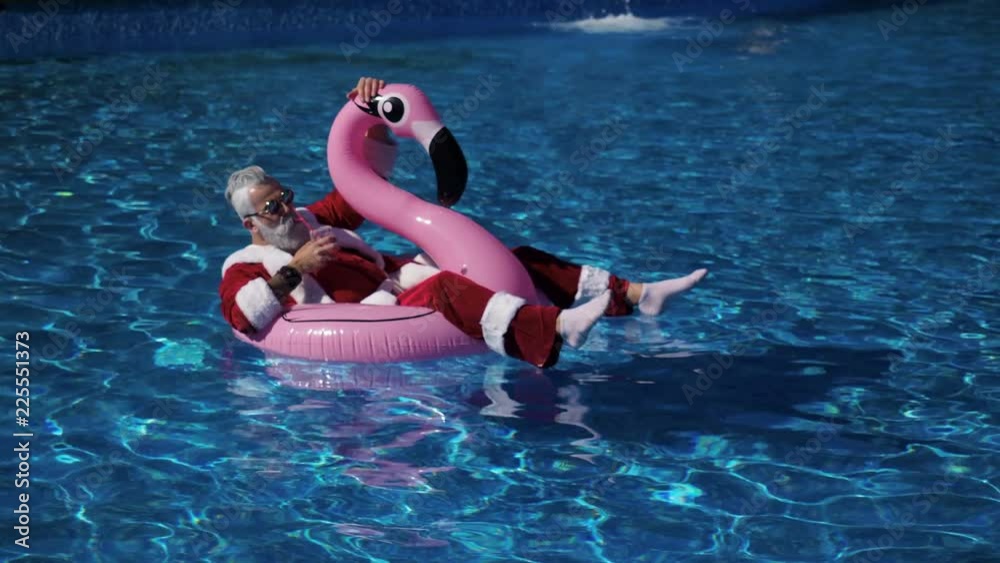 Handsome Santa floating in flamingo inflatable ring into the swimming pool with cocktail glass in hand, chilling and enjoying tropical New Year. Pleasure and happiness concept. Inspiration background
