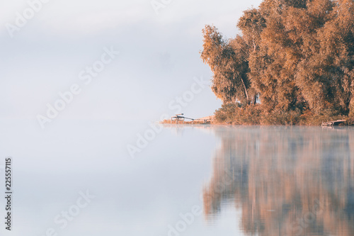 Panorama of calm autumn lake with fog over water and reflections of trees. Wooden jetty for fishing on the lake coast.