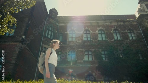 Slender Young Woman with Backpack is Walking along the Historic Building with Ivy in Summer with Nice Sun Rays and Lense Flare