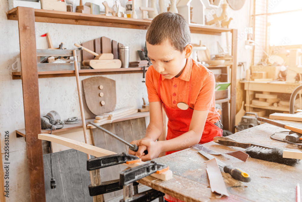 Little talented genius boy works with wood in a carpentry workshop. The concept of learning, hobbies and manual hand work for children - obrazy, fototapety, plakaty 