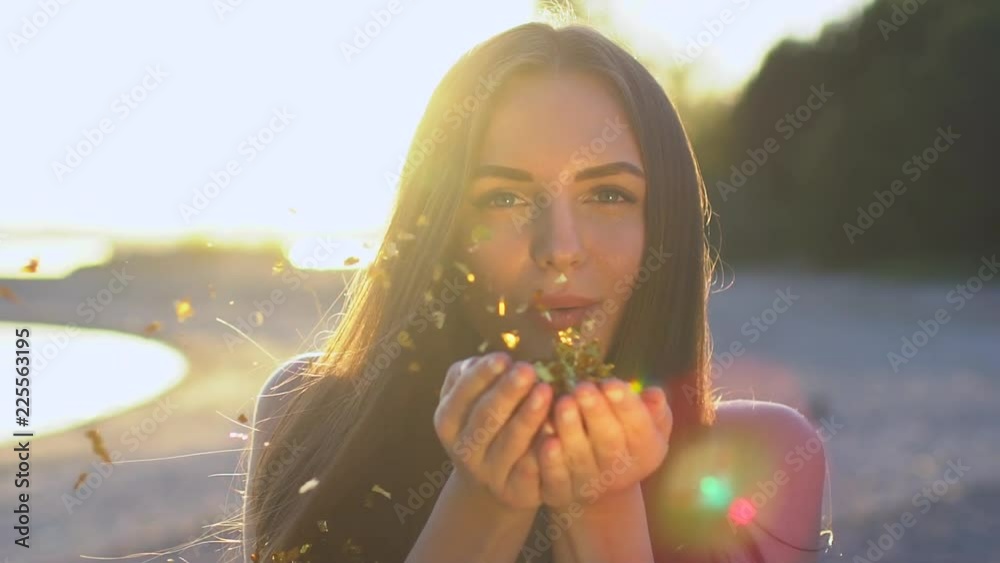 Beautiful woman blowing confetti in slow motion on the beach. Caucasian happy teenage girl blowing gold glitter off hands in summer park at sunset. Simmertime. Outside. Beauty, happiness, summer