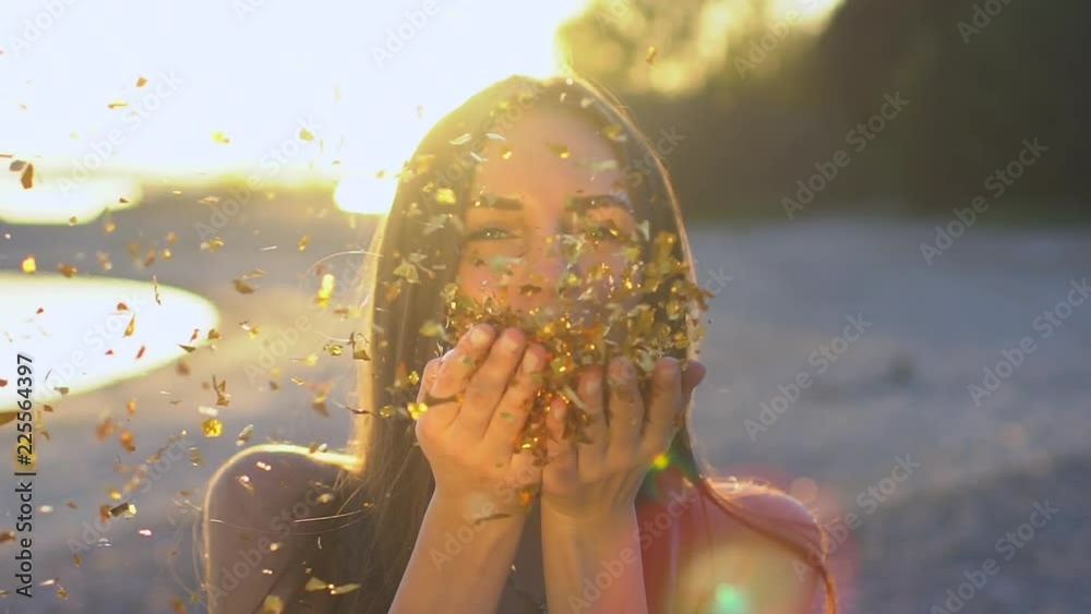 Beautiful woman blowing confetti in slow motion on the beach. Caucasian happy teenage girl blowing gold glitter off hands in summer park at sunset. Simmertime. Outside. Beauty, happiness, summer