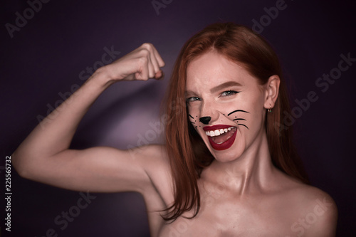 Excited happy beautiful strong redhead sports girl woman with cat carnival halloween makeup posing isolated over violet purple wall background looking camera showing biceps.