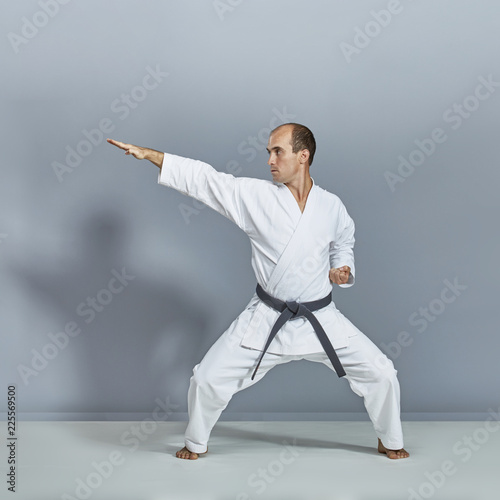 Sportsman in white karategi and with black belt is doing formal karate exercises on gray background