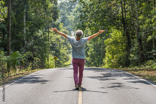 Happy Man in purple jeans and hat walking on the middle of road by yellow stripe and enjoyed surrounded jungle at sunny day. Tropical National Park with Rain Forest.