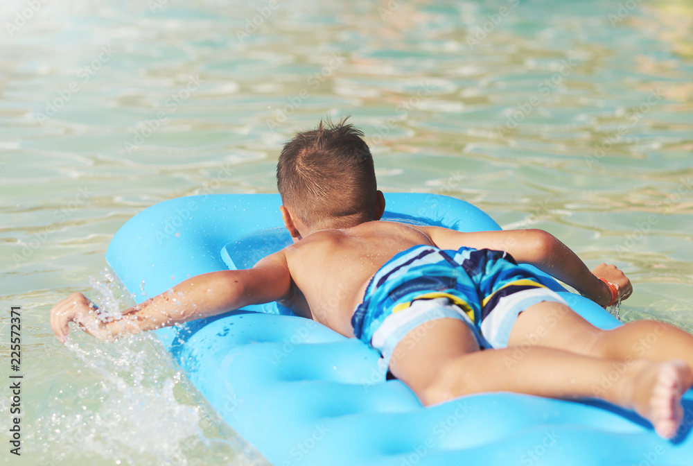 European boy is swimming in the sea on the blue air mattress, making water splashes. He is happy and enjoying his holidays. Back view...