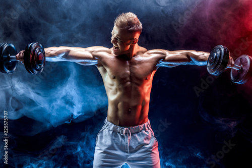Handsome bodybuilder doing Exercise for the shoulder muscles, deltoid with dumbbell. Studio shot. Smoke.