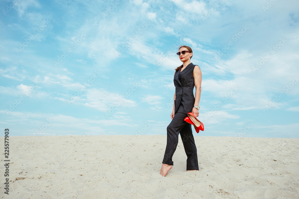 full height portrait of stylish white young woman in suit, sunglasses, red lipstick standing with red glossy high heels shoes in her hands outdoor in desert full height portrait of stylish white young woman in suit, sunglasses, red lipstick standing with red glossy high heels shoes in her hands outdoor in desert