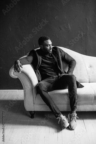 Young black man in coat on dark background sitting on a blue sofa crossed his fingers. Fashion portrait of young man in black shirt in studio. black and white portrait