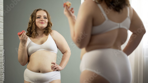 Confident fat lady in underwear happy with mirror reflection, eating sweet donut