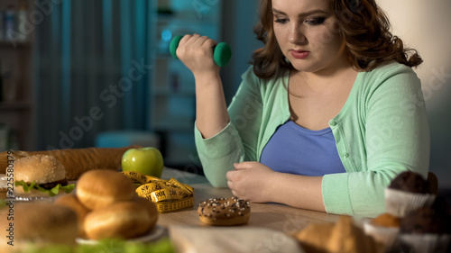 Obraz na plátně Fat lady exercising with dumbbell and looking at sweet donut, dieting willpower