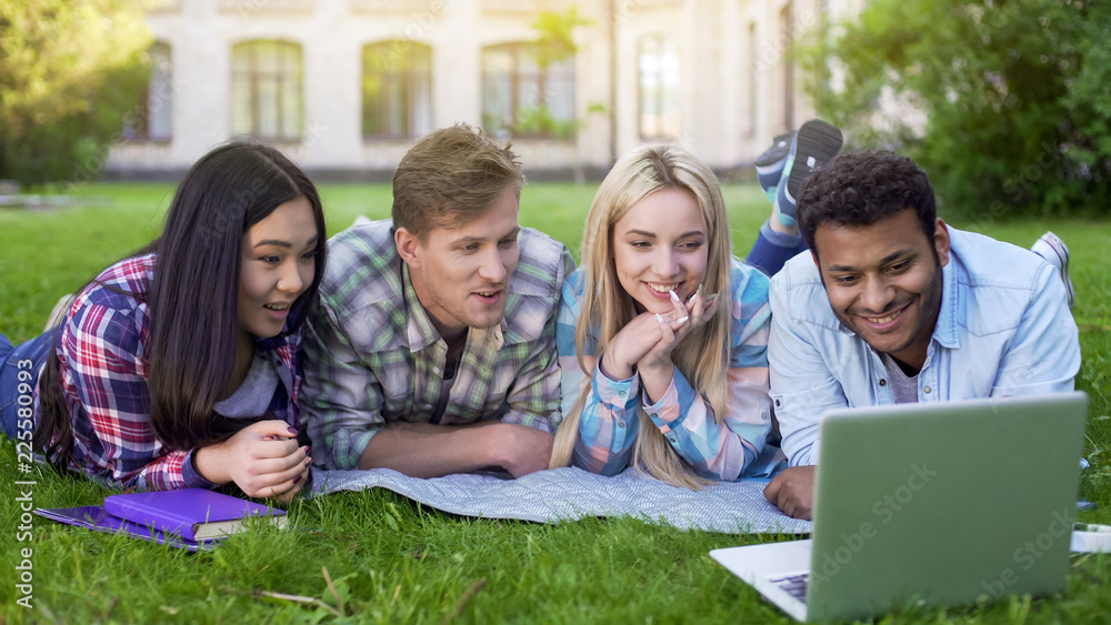Group of multi-ethnic students relaxing on grass and watching video on laptop Group of multi-ethnic students relaxing on grass and watching video on laptop