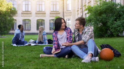 Romantic multiracial couple sitting on lawn near academy and enjoying each other