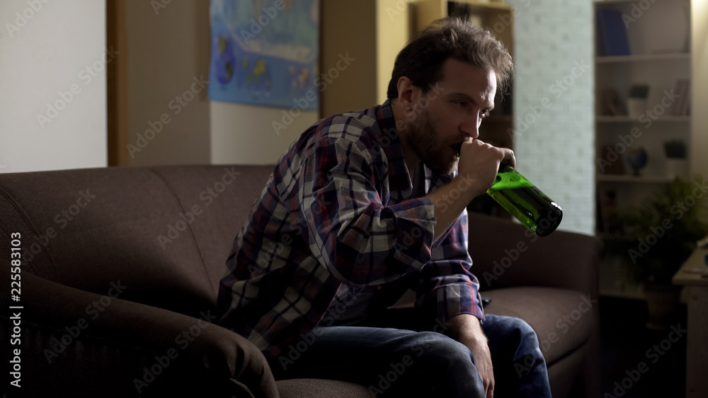 Drunken man sitting on sofa and drinking bottled beer, alcoholism and despair Drunken man sitting on sofa and drinking bottled beer, alcoholism and despair