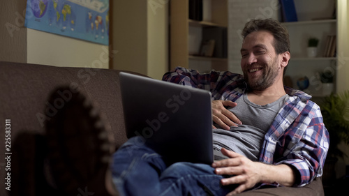 Φωτογραφία Lazy grubby male lying on couch, looking at laptop laughing at show on screen