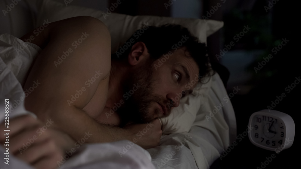 Annoyed guy in bed unable to fall asleep, looking angrily at clock, insomnia Annoyed guy in bed unable to fall asleep, looking angrily at clock, insomnia