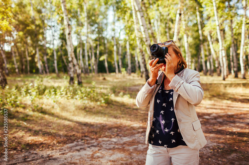 Middle-aged woman taking pictures using camera in autumn forest. Senior woman walking and enjoying hobby © maryviolet