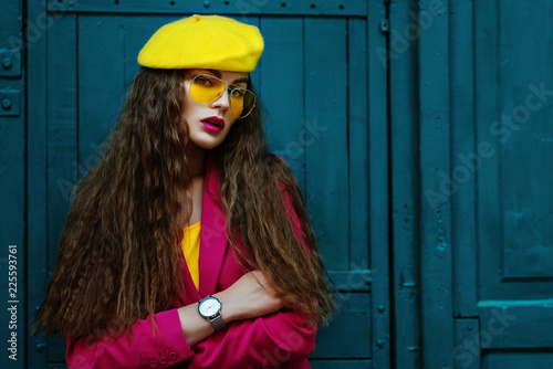 Outdoor waist up fashion portrait of young beautiful fashionable woman wearing stylish yellow beret, trendy sunglasses, pink blazer, wrist watch, posing near blue doors. Copy, empty space for text