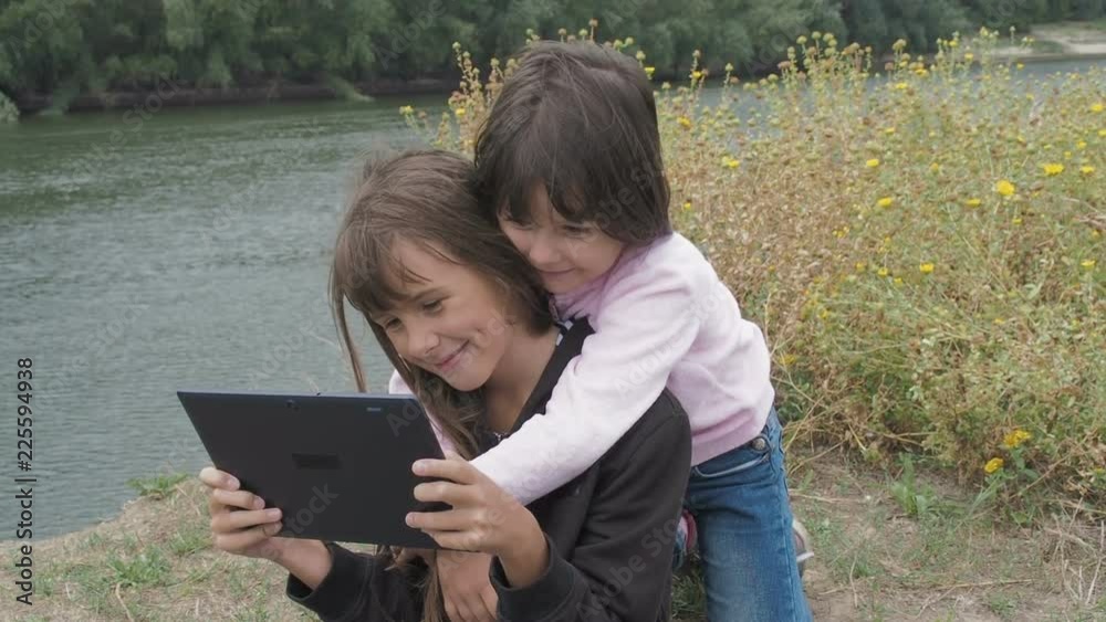 Children with a gadget in nature. Sisters play on the tablet in nature. Children with a gadget in nature. Sisters play on the tablet in nature.