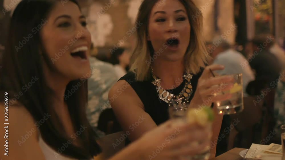 Two attractive young women talk and laugh and cheers each other over drinks at an upscale restaurant