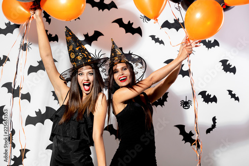 Two brunette women in black dresses and witch's hats have fun with black and orange balloons on a white background with black bats
