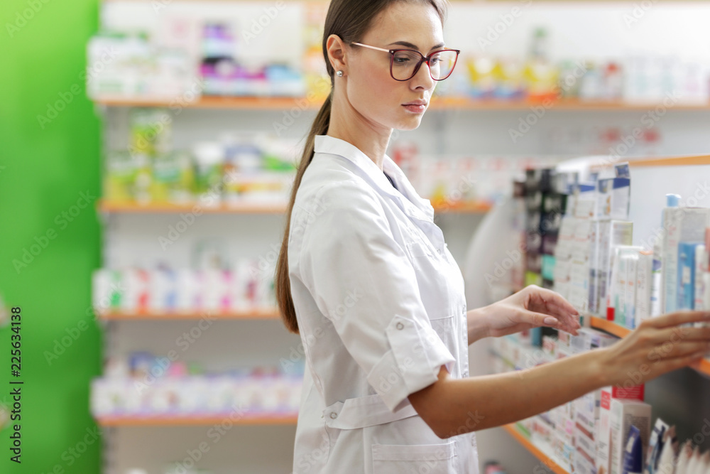 A cute slim girl with dark hair and glasses,wearing a lab coat,stands next to the shelf in a pharmacy and carefully examines it. A cute slim girl with dark hair and glasses,wearing a lab coat,stands next to the shelf in a pharmacy and carefully examines it.