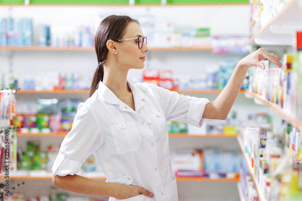 A young thin brown-haired girl with glasses,dressed in a lab coat, takes some medicines from the shelf in a pharmacy. The girl's profile is shown.