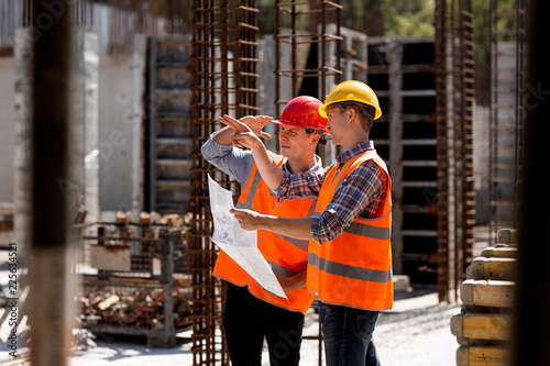 Structural engineer and construction manager in orange work vests and hard helmets explore construction documentation on the building site near the steel frames