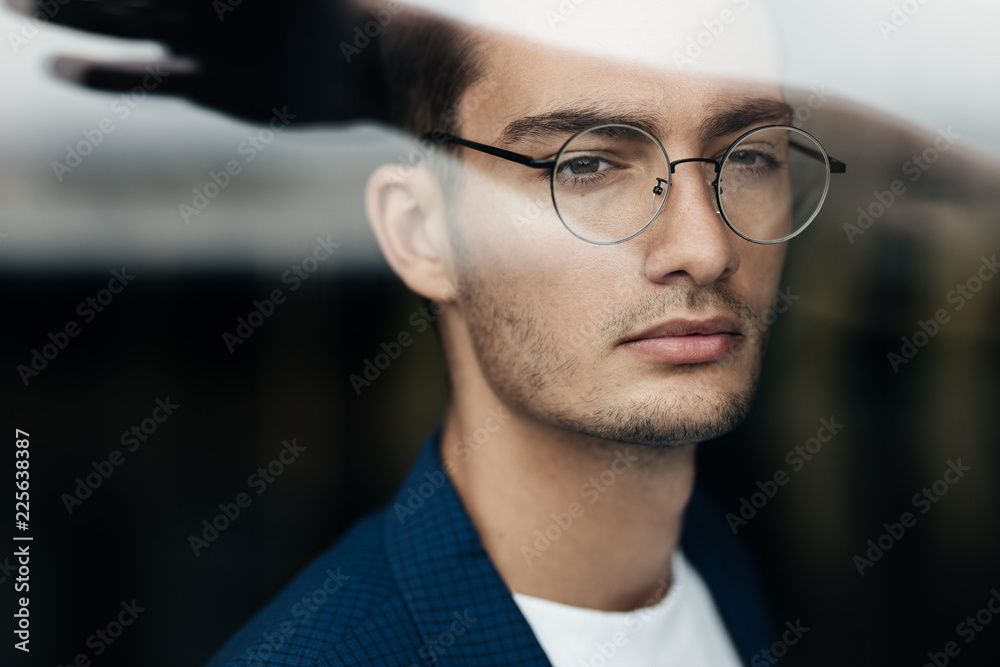 Portrait of dark-haired young architect in glasses dressed in blue checkered jacket behind the glass Portrait of dark-haired young architect in glasses dressed in blue checkered jacket behind the glass