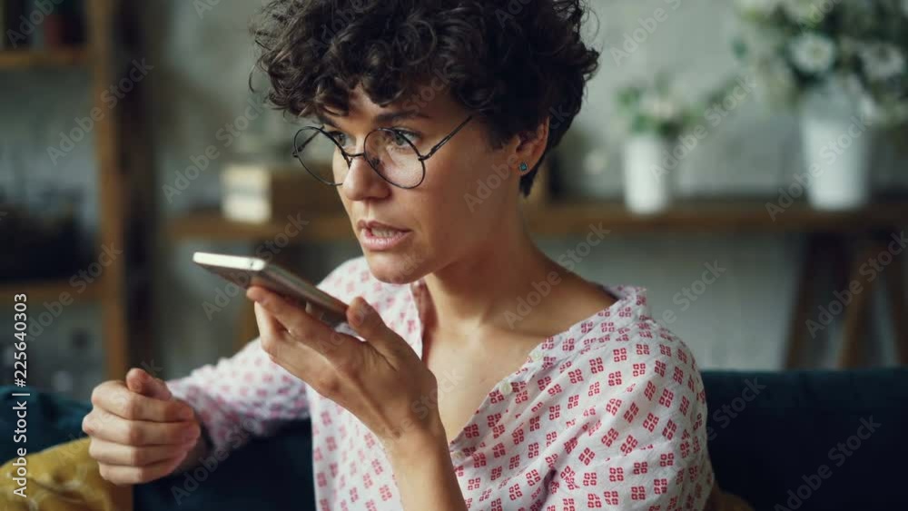 Smiling young woman is dialing telephone number then talking in loudspeaker mode holding smartphone in hand. Modern technology and communication concept.