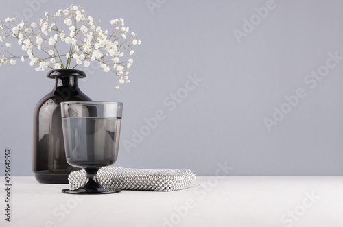 Elegant decor dressing table in minimalist style - black vase with flowers, glass, cosmetic accsssories silver bag on grey wall and white wood background. photo