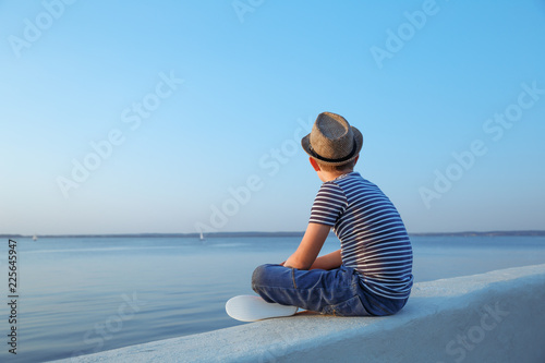 Child in marine t-shirt and straw hat sitting at the embankment and looking away at waterfront at sunset time