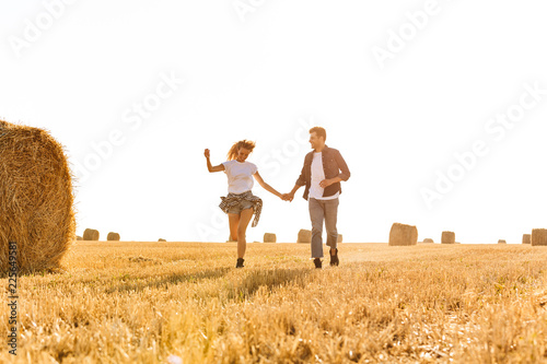 Image of beautiful man and woman running through golden field with bunch of haystacks, during sunny day